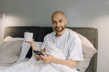 Bearded smiling man with cup of coffee in bed with phone