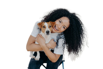 Happy curly woman tilts head, holds pedigree dog, has cheerful expression, smiles pleasantly, has...