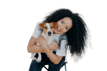 Happy curly woman tilts head, holds pedigree dog, has cheerful expression, smiles pleasantly, has curly hairstyle, wears white t shirt, isolated. Afro lady petting favourite domestic animal.