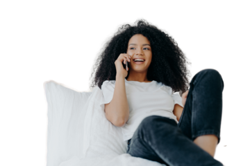 Pleasant looking curly woman has pleasant conversation, talks with friend via smartphone while relaxes in bed, has joyful expression, looks aside, wears casual clothing. People, bedding, technology