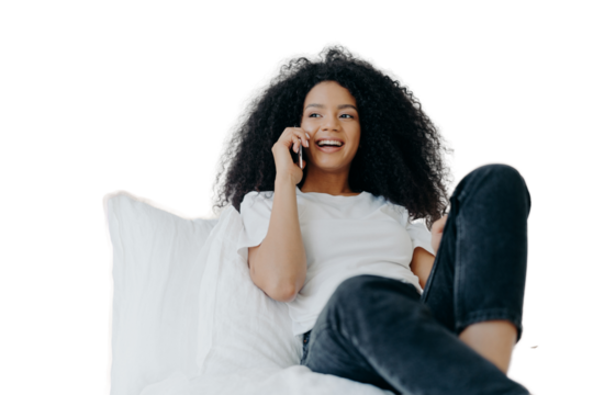 Pleasant looking curly woman has pleasant conversation, talks with friend via smartphone while relaxes in bed, has joyful expression, looks aside, wears casual clothing. People, bedding, technology