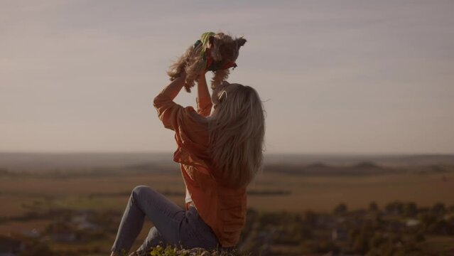 A young woman in an orange jacket sits on a mountain peak at sunset, holding and kissing and hugging her little dog on a windy day. happy watching the sunset with a beautiful view