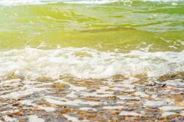 Close-up of a sea wave with white foam flowing on a pebble beach. Background with crystal clear water and sea rocks on the theme of seaside holidays and family weekends on the beach.