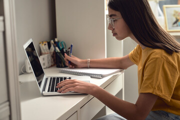 Caucasian teenage girl sitting on at the desk in bedroom and typing on laptop