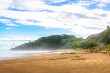 Dreamy landscape with mist over vast sand beach surrounded by lush green hills. Patches of blue sky seen through the clouds. Makorori beach, Gisborne, North Island, New Zealand