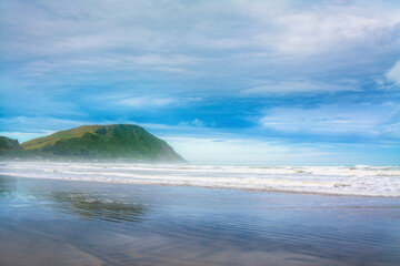 Dreamy landscape with a thick mist over ocean beach surrounded by rugged mountains. Patches of blue sky seen through the clouds. Makorori beach, Gisborne, North Island, New Zealand