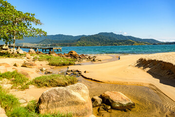 View of an empty beach, cut by a creek surrounded by rocks and a pier. small strip of sea and mountains in the background. Illhabela, Sao Paulo