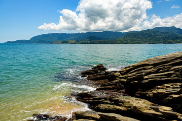 Low view of the rocks that border the beach, blue sea, mountain in the background and blue sky covered with thick white clouds. Ilhabela, Sao Paulo