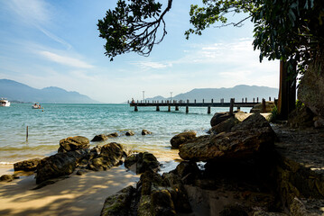 Quiet and rocky beach, in the shade under trees, blue sea and view of the wooden pier. Ilhabela, Sao Paulo
