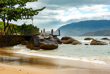 Low view of rocks on small deserted beach in cloudy day. In the background mountains covered by forest. Ilhabela, Sao Paulo