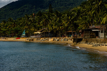 Fototapeta na wymiar rocky beach seen from the sea, fishermen's house and palm trees on the shore, mountains in the background in Ilhabela, São Paulo