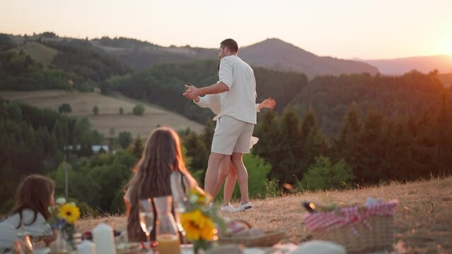 Family with children having picnic in nature, enjoying sunset. Parents dacing in front of children.