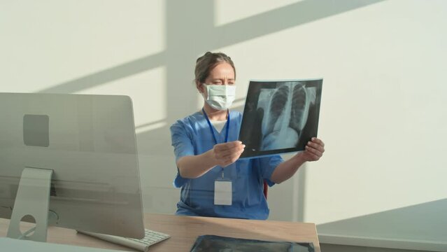 Unrecognizable female radiologist wearing blue uniform and protective mask taking seat at desk and starting work with X-ray pictures