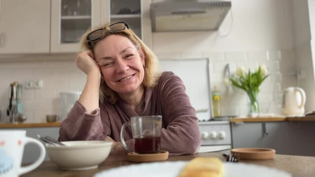 A middle-aged woman in glasses drinks coffee for breakfast sitting at the table in the kitchen sincerely laughs. Emotions