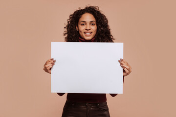Young happy smiling mixed race woman with curly hair, wears garnet turtleneck sweater, holding blank banner at studio, isolated over beige background.