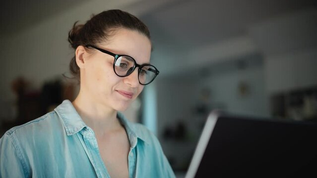 Portrait of a young adult woman wearing glasses looking to a laptop screen while working on a laptop from home.