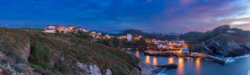Obraz premium View of the small town of Viavelez with reflections of lanterns in the harbor on the coast of Spain and where the moon breaks through the sunset clouds in the evening sky, Asturias, Spain.