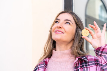 Young pretty Romanian woman holding a Bitcoin at outdoors looking up while smiling