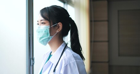 Depressed Asian young woman doctor takes off protective mask looking through the window and feeling...