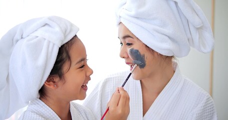 Daughter applied grey mask cream on her mom face having fun in the bedroom after shower. Family beauty treatment