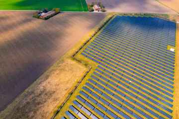 A solar park with many solar panels next to fields and a farm from drone perspective