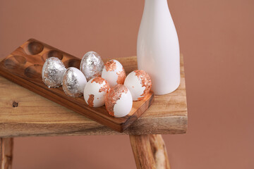 Easter decoration: eggs with silver and rose-gold metallic foil in a bamboos form on a wooden mediterranean style stool, white vase on beige colored seamless background, copy space