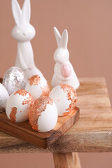Easter decoration: eggs with silver and rose-gold metallic foil in a bamboos form on a wooden mediterranean style stool, white vase on beige colored seamless background, copy space