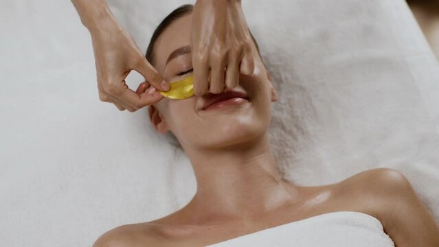 Top view portrait of young woman lying on massage bed at spa salon, beautician applying collagen patches under eyes