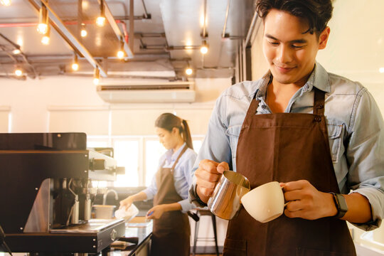 Handsome young asian barista pours milk froth into latte art coffee cup to decorate it beautifully appetizing mellow together with beautiful barista staff serving customers in the coffee cafe.