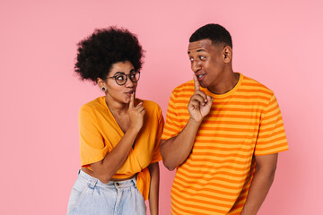 Young african couple showing silence gesture and looking at each other while standing isolated over pink background