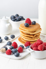 Close up of pancakes in stack with blueberries, raspberries and milk on table and white background, vertical, with copy space