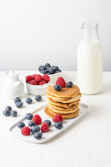 Overhead view of stacked pancakes with blueberries, raspberries and bottle of milk on table and white background, vertical, with copy space