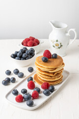 Top view of stacked pancakes with blueberries and raspberries in bowl, jar, on table and white background, selective focus, vertical