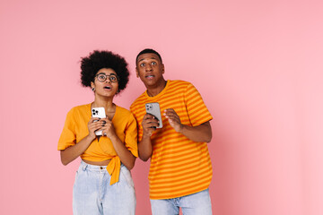 Shocked afro couple holding mobile phones and looking upward isolated