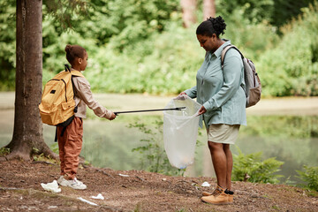 Side view portrait of black mother and daughter helping cleaning nature together and picking up trash by river, copy space  - obrazy, fototapety, plakaty