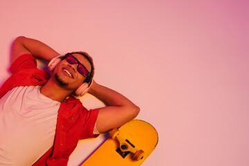 Cheerful african guy laying with skateboard isolated over gradient neon studio background