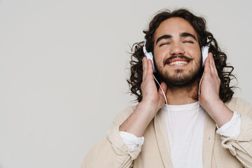 Smiling hispanic man listening music with headphones while standing with eyes closed isolated over...