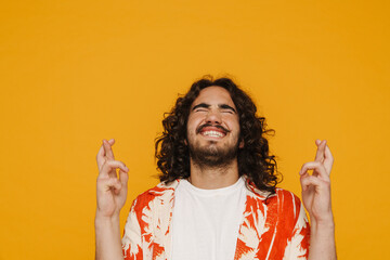 Cheerful hispanic man holding fingers crossed for good luck while standing isolated over yellow wall