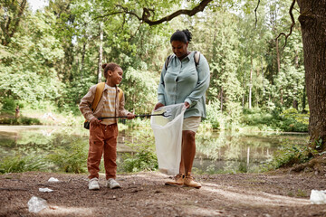 Full length portrait of black mother and daughter helping clean nature and picking plastic bottles in forest - obrazy, fototapety, plakaty