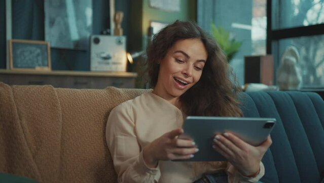Footage of good-looking young relaxed woman sitting with digital tablet in hands, browsing through device. Cute girl watching funny video and chatting with friends in comfortable modern living room