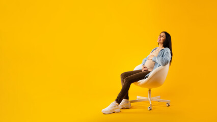 Relaxed pregnant woman sitting in chair and caressing belly, happy expectant lady looking at free...