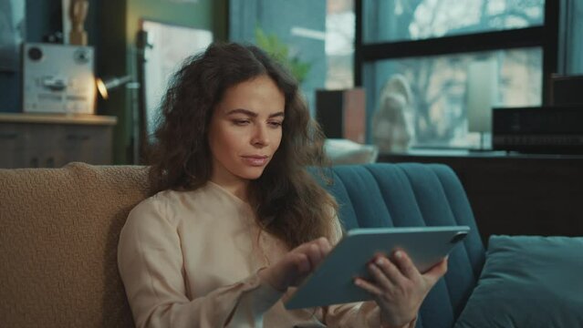 Footage of good-looking young relaxed Caucasian woman holding digital tablet in hands, connecting with others, posting on social media. Charming girl with long hair smiling at camera in cozy apartment