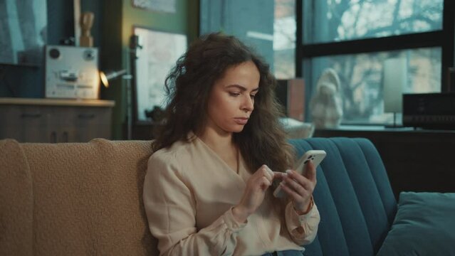 Footage of good-looking focused young Caucasian woman in elegant clothes scrolling through social media feed, checking messages on smartphone. Smiling girl with gadget sitting on comfortable sofa in