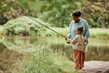 Side view portrait of caring black mother teaching little girl fishing by beautiful forest lake, copy space  - obrazy, fototapety, plakaty