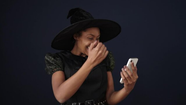 African american woman wearing witch costume using smartphone over isolated black background