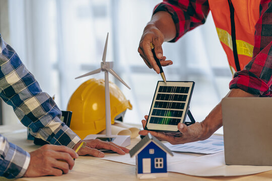 Construction team starts new project plans behind yellow helmets on table in office to discuss renewable energy construction projects with wind turbines and solar cells and houses on table.