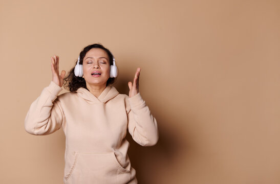 Relaxed delightful multi-ethnic middle-aged woman with her closed eyes, wearing beige hoodie and wireless headphones, sings, listens to music, enjoying new sound track, on isolated cream background