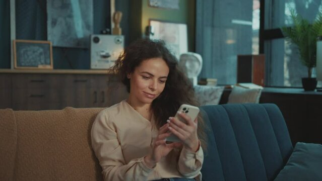 Portrait of attractive joyful Caucasian young woman chatting in messenger. Beautiful girl sitting on couch and typing messages on screen of smartphone in cozy home. Indoors. Daytime