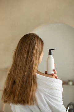 Woman holds a white bottle of cosmetics in her hand, standing with her back to the camera. Mock up shampoo, gel.