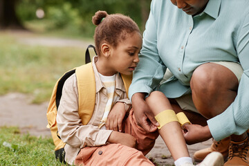 Portrait of black little girl injured in nature hike with mother putting bandage on knee outdoors
